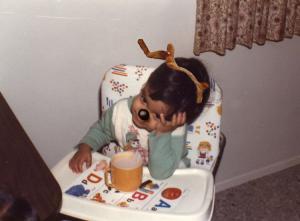 A baby, with antlers and black deer nose, sits in a highchair with a cup of tea.