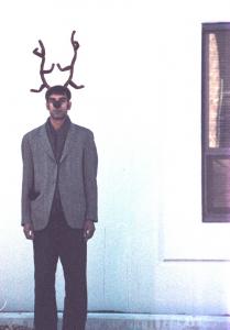 A man, with antlers and deer nose, stands in front of a white wall looking quite serious.
