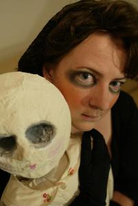 Close-up of a woman's face. Her eyes look sunken in, thanks to heavy makeup. Next to her face is a large creepy doll's face. Its eye sockets are empty in its white round head.