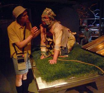 A man dressed in rags sits like a dog on a platform covered in artificial grass. A man wearing a bucket hat and carrying a tape recorder talks with him.