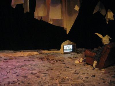 A large rabbit wearing a ratty robe and bunny slippers slumps in a lazy boy recliner watching television. The floor is covered with newspaper and objects covered in sheets hang from the ceiling.