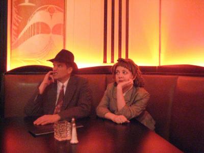 Two 1940’s detectives sit in a booth at an art deco bar looking bored.