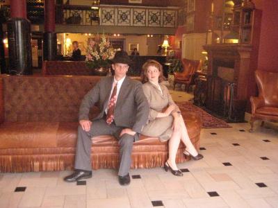 A pair of 1940s detectives sit in the lobby of an art deco hotel looking at each other puzzled.