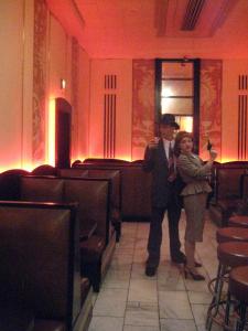 1940’s detectives stand in the middle of an art deco hotel. One of them is holding her gun up