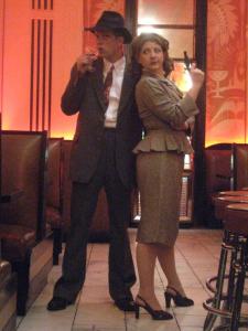 1940’s detectives stand in the middle of an art deco bar. The man drinks a martini while the woman holds a gun up and looks on disapprovingly.