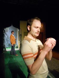 A mustachioed man in a tan sweater looks quizzically at a Swiss Army knife. Behind him, another man in bell bottoms and a leather jacket stands framed by an archway. There is astroturf and some greenery.