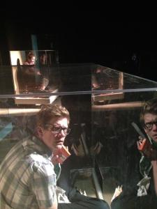 A man with a plaid shirt and glasses looks confused. He is in a large plexiglass box and his reflection is shining in the plastic wall. In the distance, another woman sits in a smaller plexiglass box.