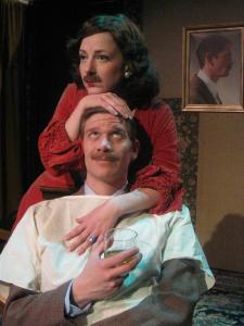 A man with a large mustache, dressed in a suit, and wearing a hospital gown sits holding a glass of whiskey. A woman, wearing a red velvet gown, pearl earrings and the man’s mustache stands behind him. The woman embraces the man’s head and looks dispassionately into the distance. Behind the couple a portrait of the man in profile can be seen.