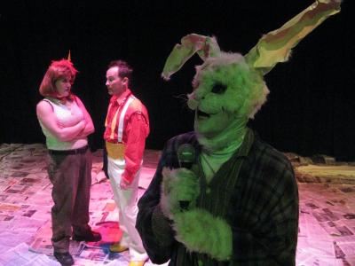A large rabbit wearing a ratty robe and bunny slippers sings into a microphone. Behind him a magician with one arm speaks to his assistant. His assistant has the legs of a mechanic.