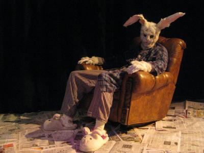 A large rabbit wearing a ratty robe and bunny slippers slumps in a lazy boy recliner. The floor is covered in newspaper.