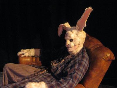 Closeup of a large rabbit wearing a ratty robe and bunny slippers slumped in a lazy boy recliner. The rabbit looks at the camera menacingly.