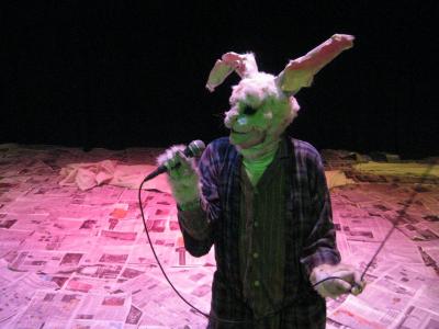 A large rabbit wearing a ratty robe and bunny slippers stands on a floor covered with newspapers. The rabbit sings into a microphone.