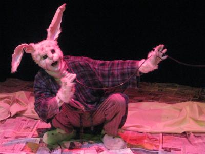 A large rabbit wearing a ratty robe and bunny slippers squats on a floor covered with newspapers. The rabbit holds a microphone and sings.
