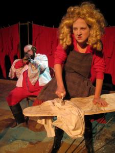 A woman in an extravagant blond wig angrily irons a white shirt on a white ironing board. Behind her, a larger man in red long underwear sits with a rifle. The floor is covered in mud and a clothing line of red long underwear hangs in the background.