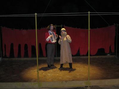 Two people stand in a shallow mud pit. One is tall with a large wig. The other wears a pith helmet and a long coat. They are in a spot light. A clothing line of red long underwear hangs in the background.