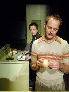 A mustachioed man talks while looking at an onion. Behind him, a woman with short, choppy hair stands at a stove glaring at the man.