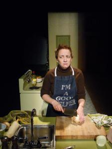 A woman with short, choppy hair is cutting onions next to a sink. She wears an apron that says “I'm a good helper” and she looks miserable.