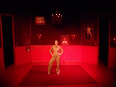 A woman strikes a strong pose, arms akimbo, center stage. The room she stands in is entirely red and decorated with her portraits.