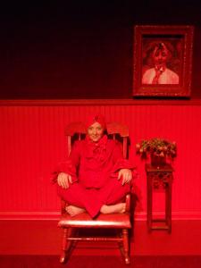 A woman sits in a large red rocking chair and smiles at the camera. She’s dressed entirely in red. Everything in the room is red. Her portrait, painted in red, hangs on the wall.