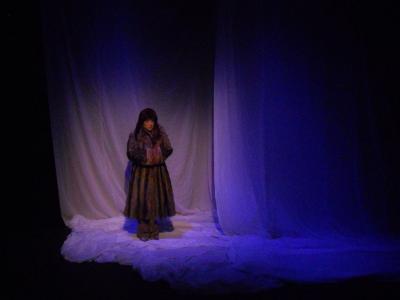A woman dressed from head to toe in furs stands on a stage draped in white fabric. She looks cold.
