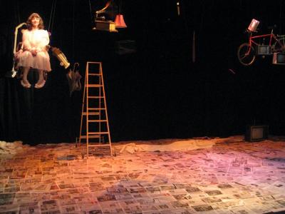 A wooden ladder is set up in the middle of an empty stage covered with newspaper. Suspended from the ceiling are various objects including a woman in a rocking chair.