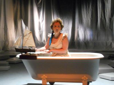Mary Shelley is sitting up in her bathtub writing on a writing desk built for her bathtub. She has small sail boat sitting on her desk and she looks worried.