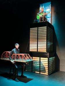 A person is sitting on top of a tall structure that represents the Sears Tower.  They are playing a green guitar and singing. Below the tower sits a person wearing all black with a bridge built around their waist.