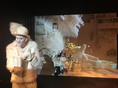 A man dressed all in white with a fur coat and hat with a large feather in it smiles creepily. He is holding a large white container. Behind him, on the wall, is a projection of his own profile. In the distance, with the projection on her, is a woman covered in stuffed animals. She is waving.