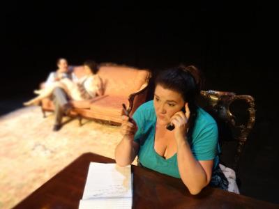 A down shot of a woman sitting at a desk, talking on the phone. She has a pad of paper in front of her. Behind, out-of-focus, in the distance, a woman and a man sit on a pink couch. She has her legs crossed over his.