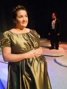 In the foreground, a very pregnant woman in a long green period dress, holds her belly. In the background, A man dressed as Lord Byron stands looking off into the distance.
