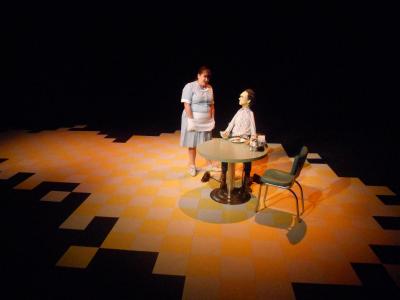 A life-size puppet of Tommy Lee Jones sits at a diner table that is surrounded by a yellow and white checkered floor surrounded by darkness. A waitress stands next to the table as if she is about to take his order.