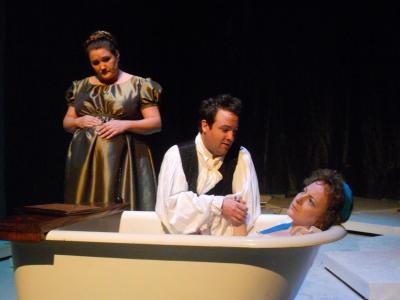 A woman, looking upset, lays in a bathtub and holds the hand of a man kneeling behind the tub. Standing in the background is a pregnant woman in a green dress. They are all in period clothing.