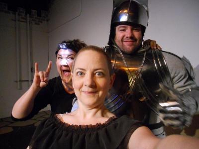 Two women, and a man dressed in a suit of armor poise for a selfie. One of the woman is wearing a head lamp and making the devil horns rock-and-roll sign with her hands.