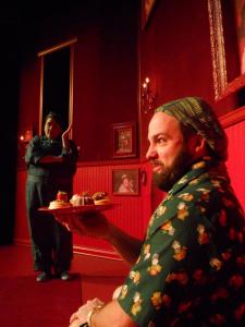 In the foreground a man contemplates a plate of pastries. A woman dressed in work coveralls looms in the background. The room is well decorated and entirely red.