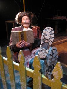 A person with a large brown wig and mustache sits reading a book. Their dirty boots are propped up on a muddy pea green wooden fence.