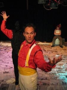 A magician and his disembodied arm strike a pose. A woman dressed as the magician’s assistant but wearing mechanic’s pants sits on a television looking angry. A red bicycle and two televisions hover above her.