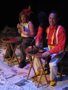 A magician and his disembodied arm enjoy a t.v. dinner on a small tv table outfitted with a microphone. The magician’s assistant sits at a t.v. table next to him examining a boxed frozen dinner.