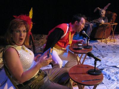 A one armed magician sings into a microphone at a small t.v. table. A large rabbit slumps in a lazy boy recliner behind him. In the foreground the magician’s assistant looks shocked.