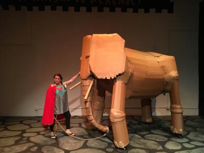 A woman in an inexpensive looking knight costume stands next to a life size cardboard elephant.