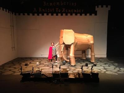 A woman in an inexpensive looking knight costume stands next to a life size cardboard elephant. In front of her there is a row of overhead projectors and on the wall behind there is an outline of a castle and the title "Remembering a Knight To Remeber."