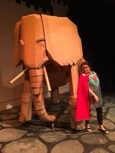 A woman in an inexpensive looking knight costume leans against a life size cardboard elephant.