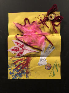 A colorful collage on a bright yellow piece of paper. Bright pink feather, red yarn, and strips of white paper are glued on. There are also some green doodles and red-and-blue sponge shapes stamped on in paint.