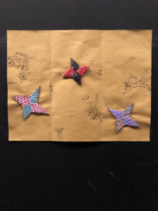 Three colorful origami stars are pasted to a yellow page. Surrounding them are pen-and-ink doodles of galloping horses, cowboy boots, and a carriage that has lost a wheel.