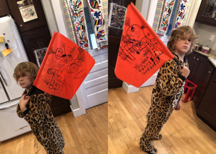 A child stands in their kitchen wearing a leopard onesie and carrying a homemade, red, BBPSS flag.