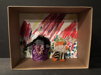 A diorama in a small cardboard box. The background is colorful and squiggly with an orange butterfly sticker. Standing in front are two crocheted characters. One has long black hair, a purply muumuu and a flower necklace. The other has short yellow hair, striped pants, a white top, and a gold chain.
