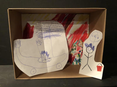 A diorama in a small cardboard box. The background is colorful and squiggly with butterfly stickers. Leaning on one side is a pen-and-ink chaotic drawing of a panicked person driving a banana car. On the other side, is a stick figure next to a red bucket.