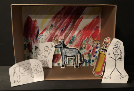 .A diorama in a small cardboard box. The background is colorful and squiggly with butterfly stickers. Leaning against the back is a black-and-white drawing of two people dancing. Next to them is a cutout painting of a donkey. Leaning on either side of the box is a drawing of an Eeyore-type creature and a drawing of a partying stick figure.