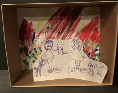 A diorama in a small cardboard box. The background is colorful and squiggly with butterfly stickers. In front is a pen-and-ink drawing of two people clinking wine glasses. They each have a dog next to them.