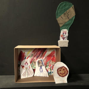 A diorama in a small cardboard box. The background is colorful and squiggly with butterfly stickers. Leaning against the back are collages of two odd creatures. Leaning against the right side of the box is yet another creature that looks like it has a smiling pepperoni pizza face. Above the box is a hot air balloon with a fourth creature in the basket.