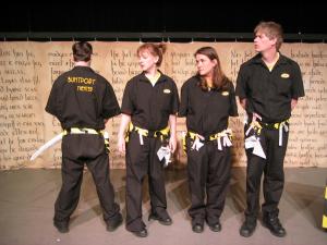 A group of four dead pan looking people, wearing utility suits and utility belts with origami paper props hanging off of them, are standing in a line. One of the people is facing backward and on the back of their utility suit are the words Buntport Theater embroidered in yellow.
